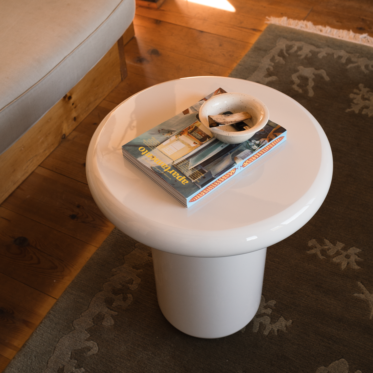 Mieux round mushroom shaped side table in a cream high gloss, styled with a magazine and a small white dish in a cozy room setting with wooden floors and a patterned rug.