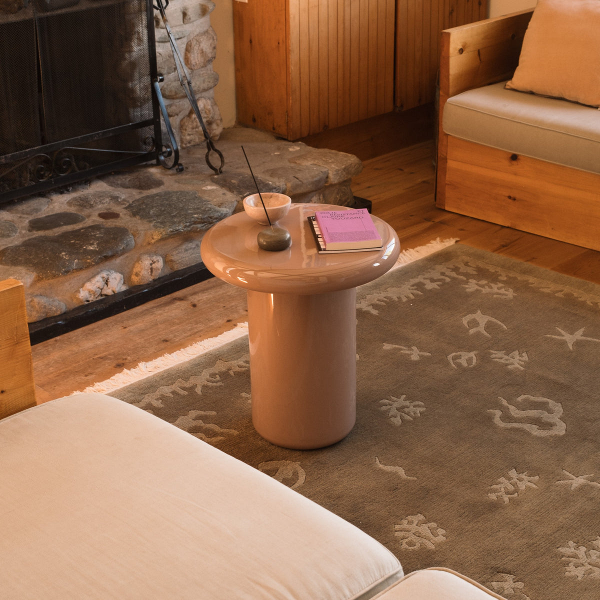 Mieux mushroom shaped glossy side table in a dusty pink, styled in a cozy living room setting next to a stone fireplace and wooden furniture.