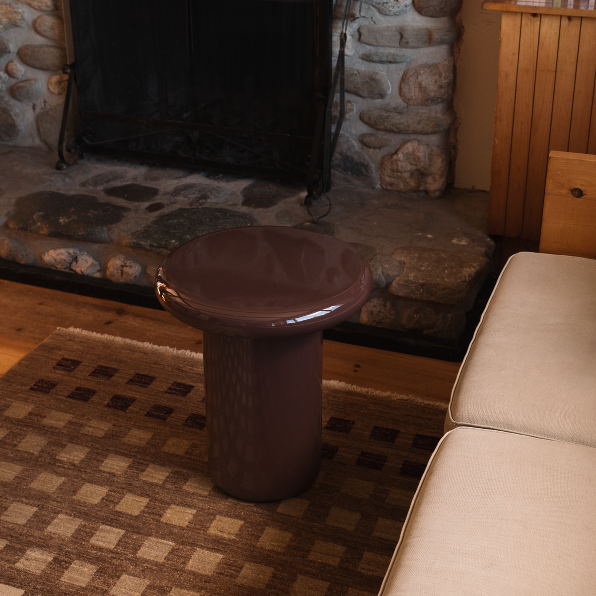 Mieux glossy, round side table in dark red, positioned in a cozy living room setting next to a light beige sofa and in front of a rustic stone fireplace.