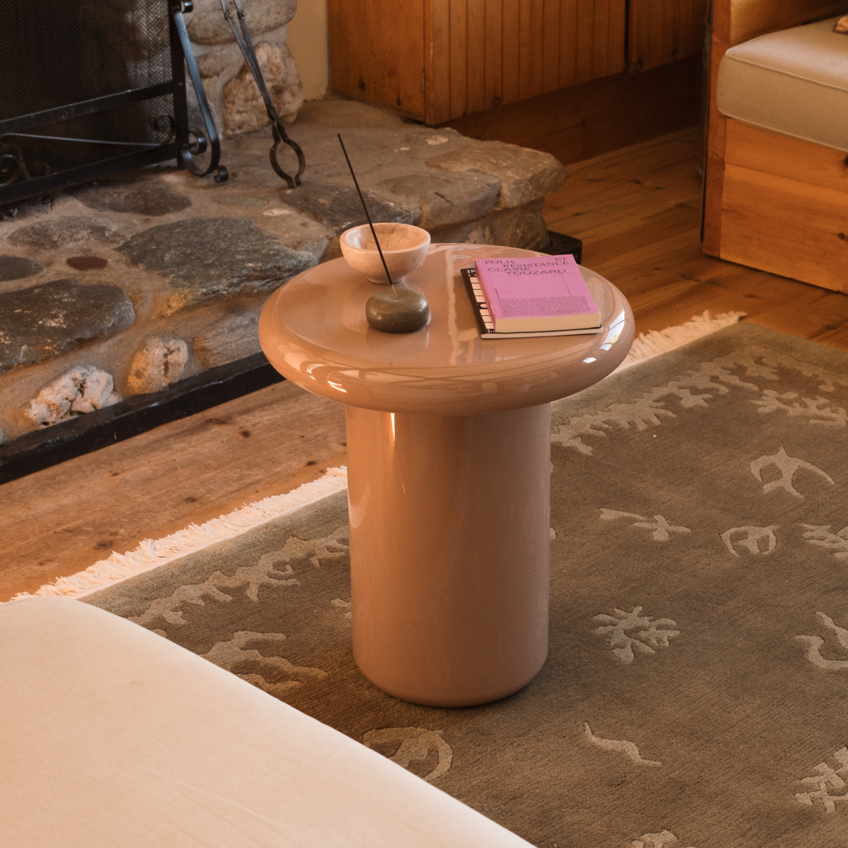 Mieux mushroom shaped glossy side table in dusty pink, styled in a cozy living room setting with a patterned rug, fireplace, and wooden furniture.