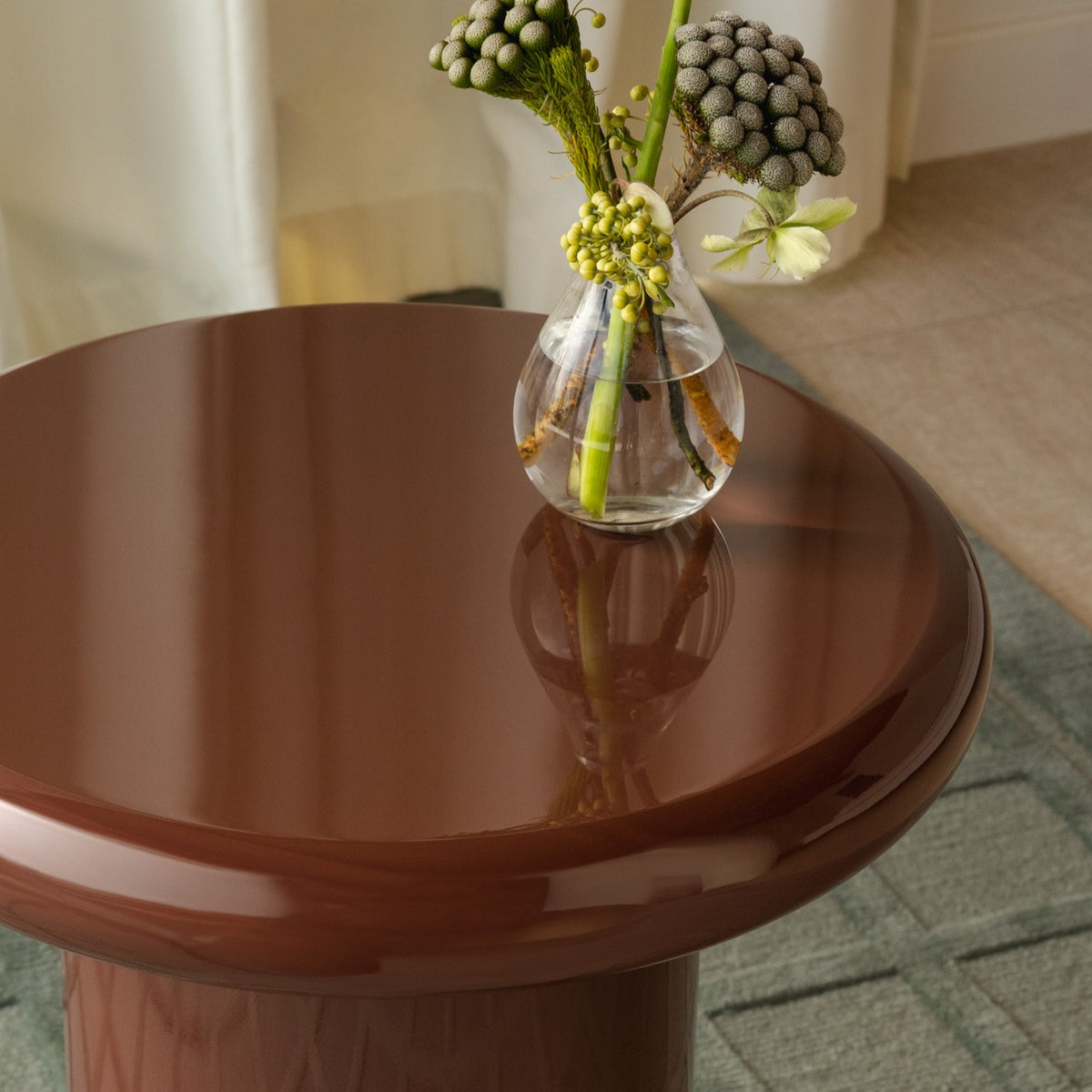 Mieux glossy round side table in deep burgundy styled with a small clear glass vase holding green stems and dried pods, set in an indoor living room with a rug and curtains visible.