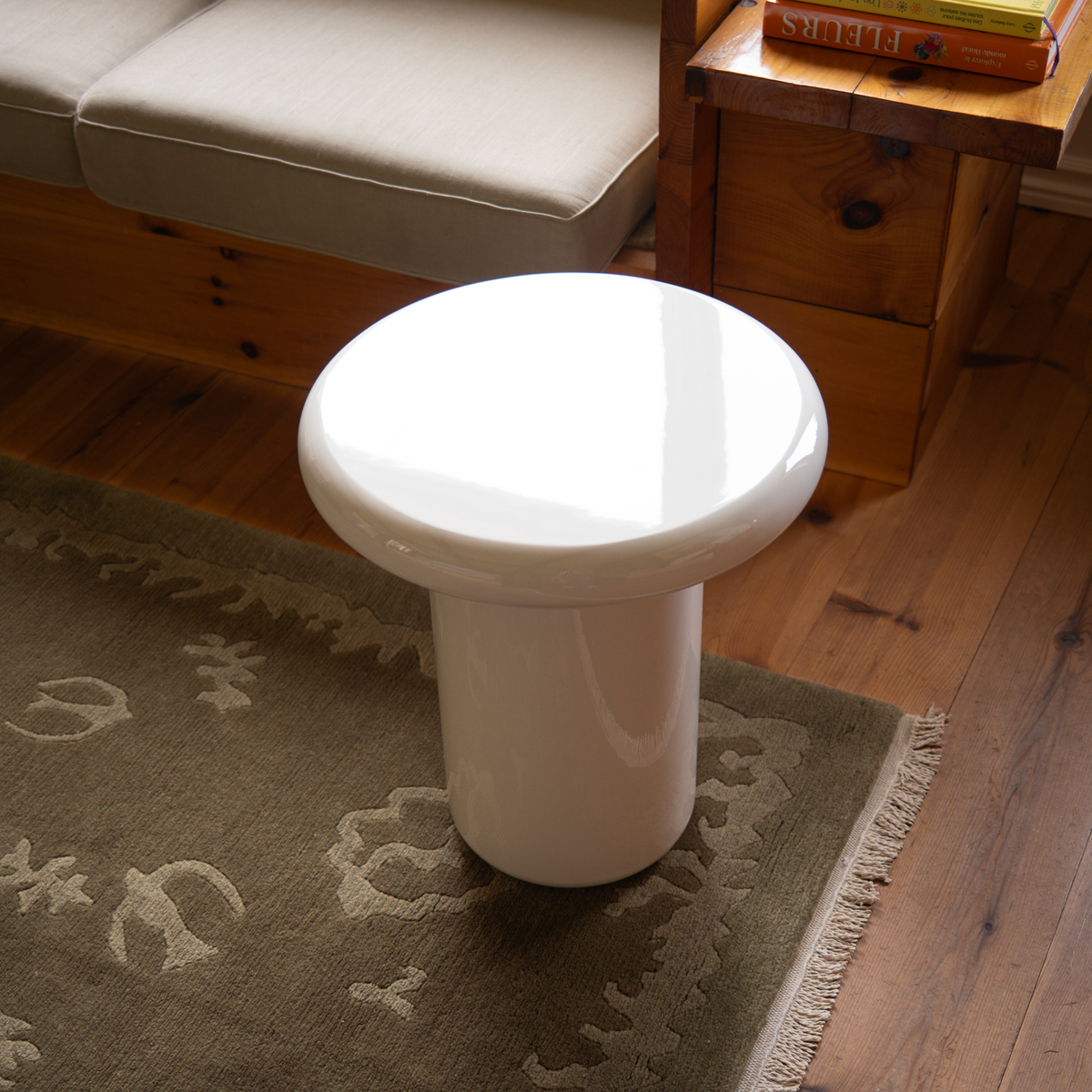 Mieux glossy cream side table with a mushroom shape, shown in a home setting on a patterned rug next to a wooden bench and side table.