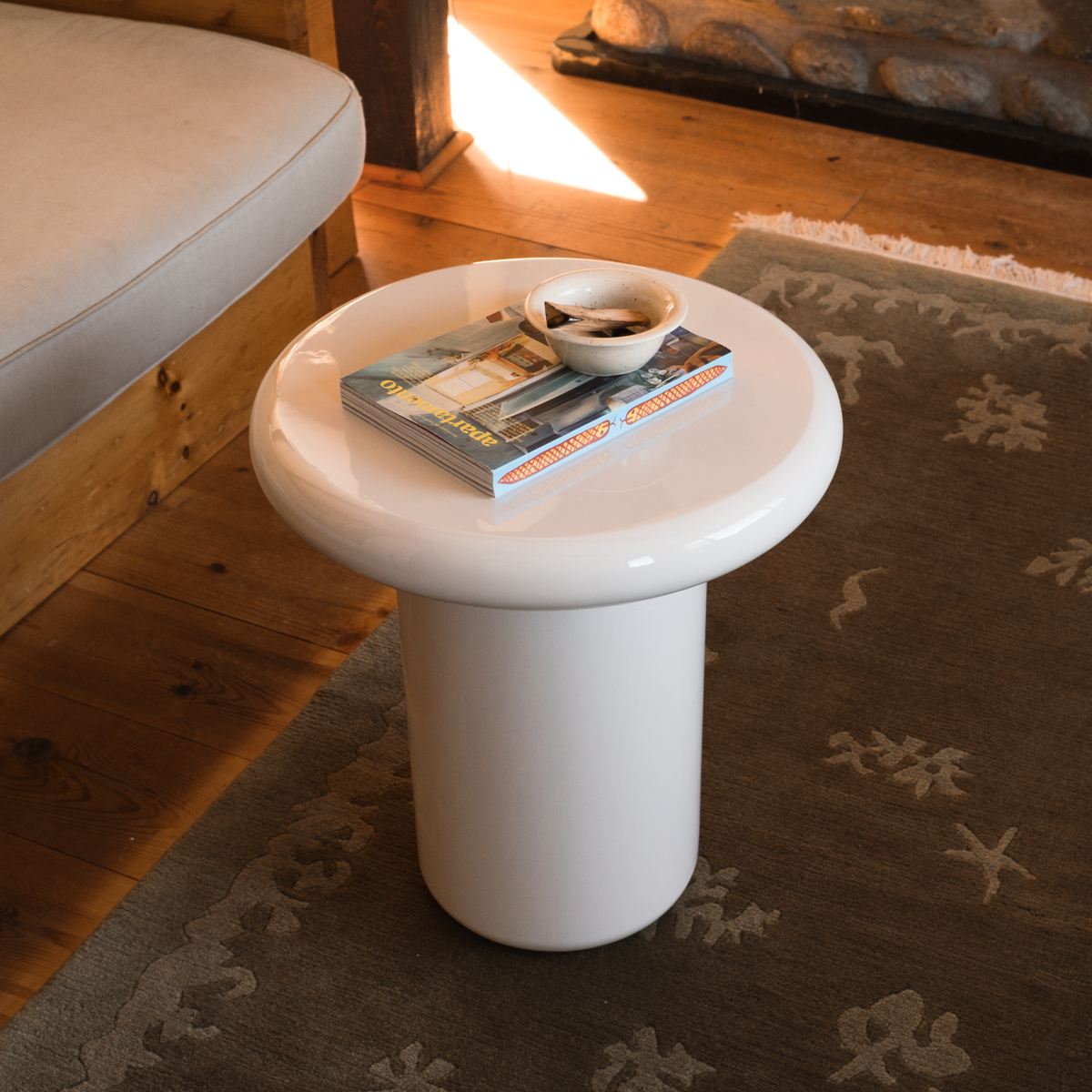 Mieux mushroom shaped glossy side table in cream, styled with a magazine and a small bowl in a warm, mid century living room setting.