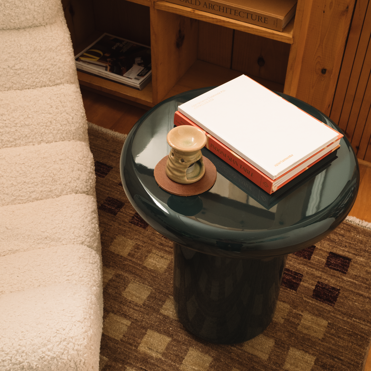 Mieux round dark teal glossy side table in a cozy room setting, styled with a stack of books and a small ceramic cup on a patterned rug.