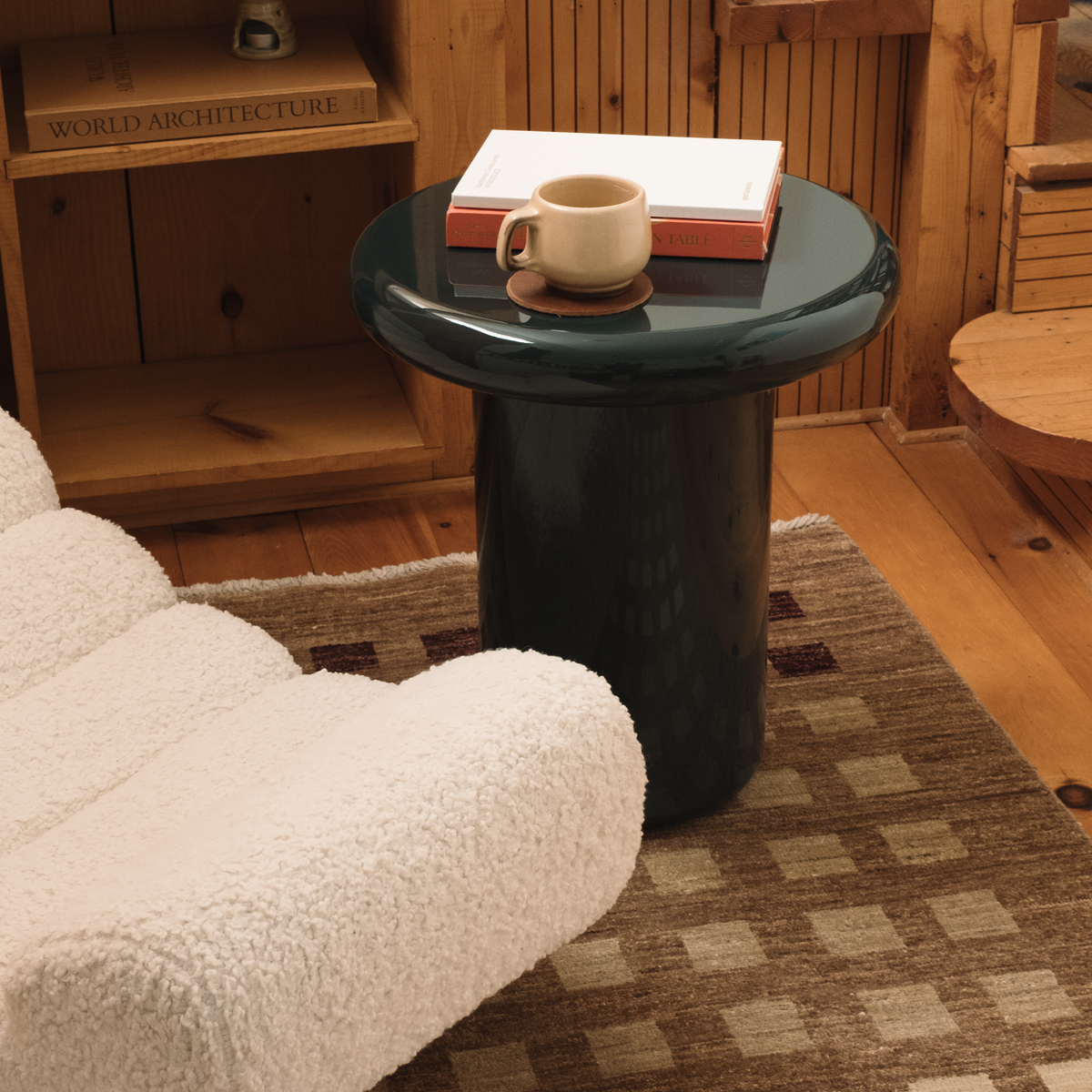 Mieux round side table in a glossy dark teal, styled in a cozy room setting next to a white boucle chair with books and a mug on top.