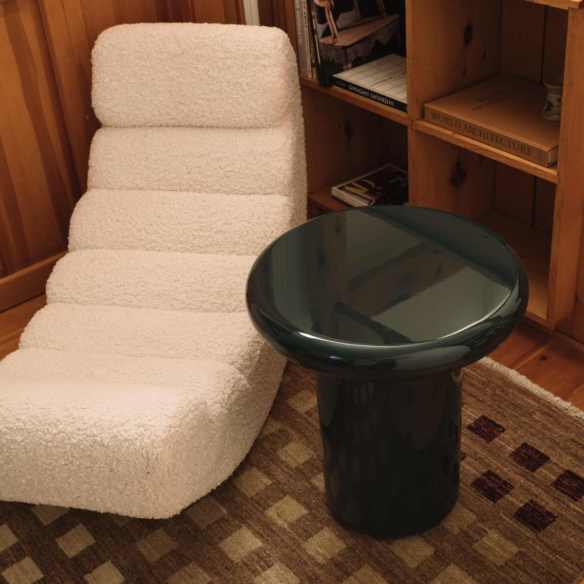 Mieux glossy mushroom shaped side table in dark teal, positioned next to a cream bouclé lounge chair in a warm wood-paneled room.