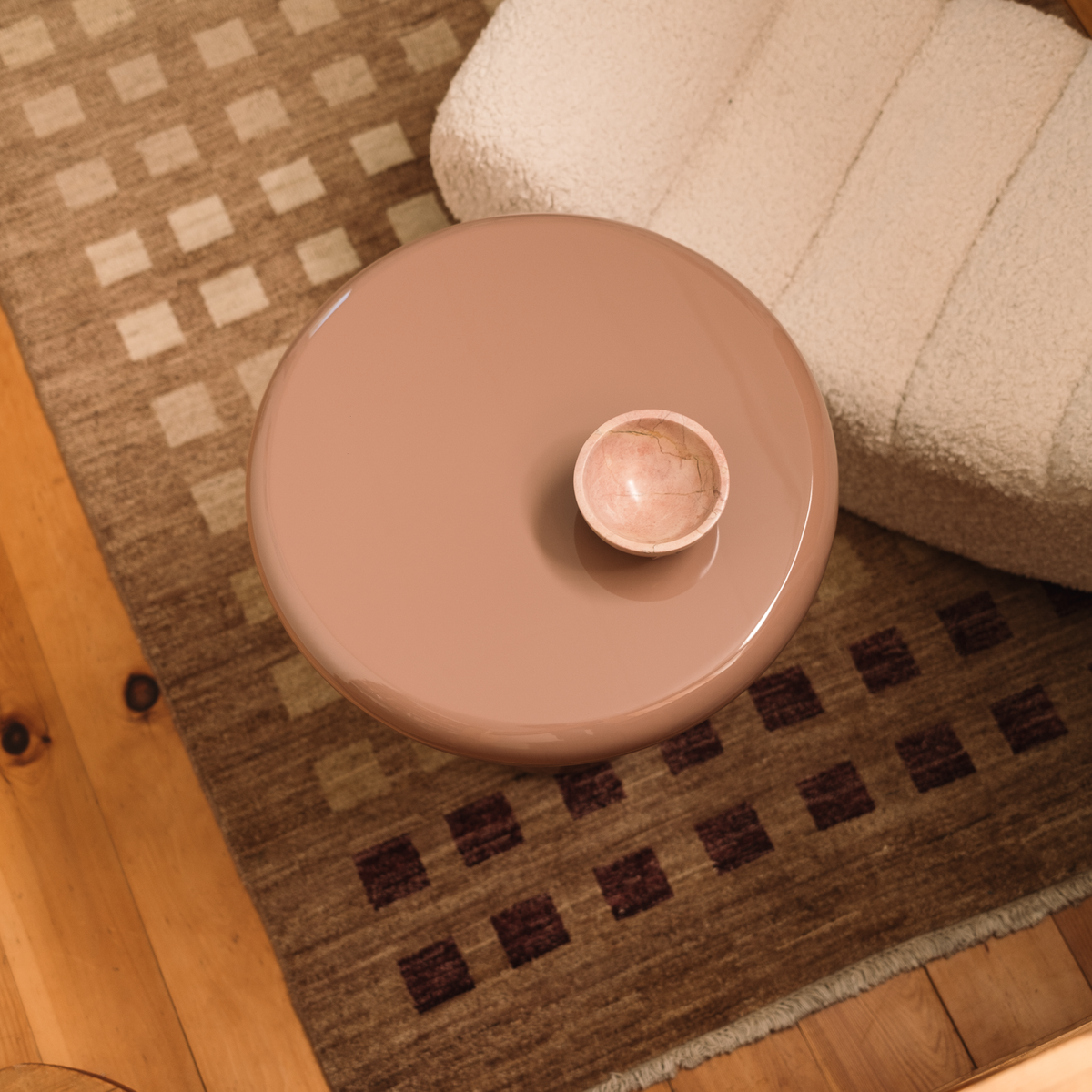 Mieux round, high gloss side table in a dusty pink finish, featuring a small matching ceramic cup on top. The table is styled in a cozy room setting on a patterned brown rug next to a cream-colored textured pouf.