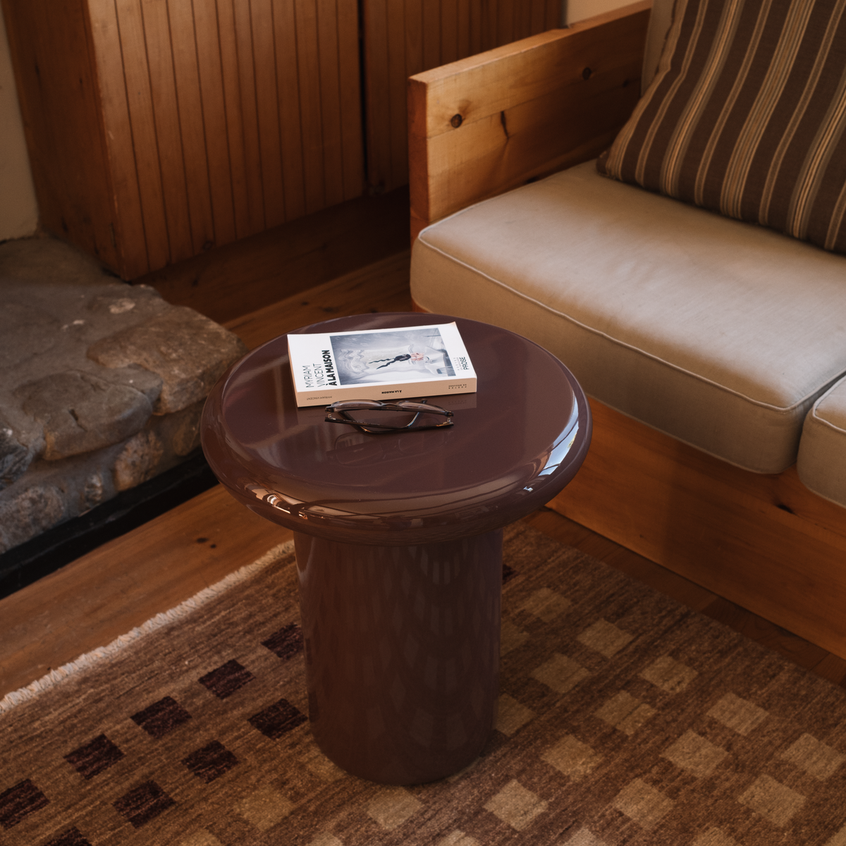 Mieux mushroom shaped high gloss side table in dark red, styled in a cozy living room next to a wooden sofa with beige cushions.